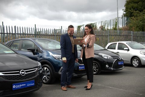 A man and a women standing in front of a car and smiling while looking at the Ipad